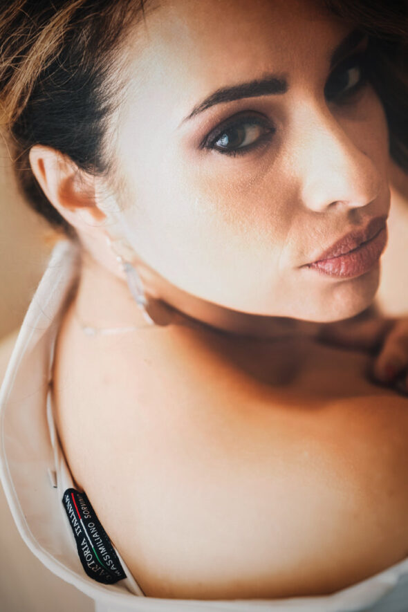 Ritratto di una donna con trucco elegante e orecchini, sguardo intenso e capelli raccolti. La luce mette in risalto la sua pelle e i dettagli del vestito.