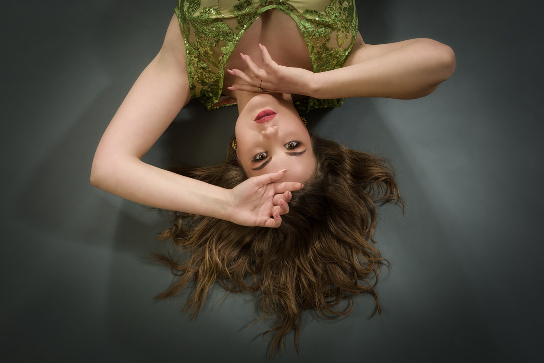 Modella con capelli mossi e abito verde, sdraiata su un fondo grigio, in una posa artistica e contemplativa.