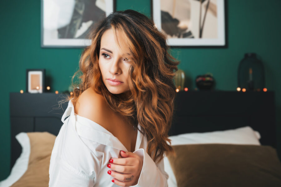 Modella con capelli ricci e lunghi, vestita con una camicia bianca, posa in una camera elegante con pareti verde scuro e decorazioni moderne.