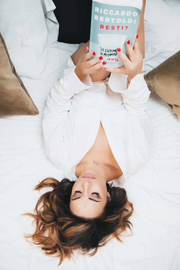 Donna sdraiata su un letto bianco, indossando una camicia bianca, mentre tiene un libro tra le mani e guarda verso l'alto, con un'espressione serena. Cuscini beige sullo sfondo.