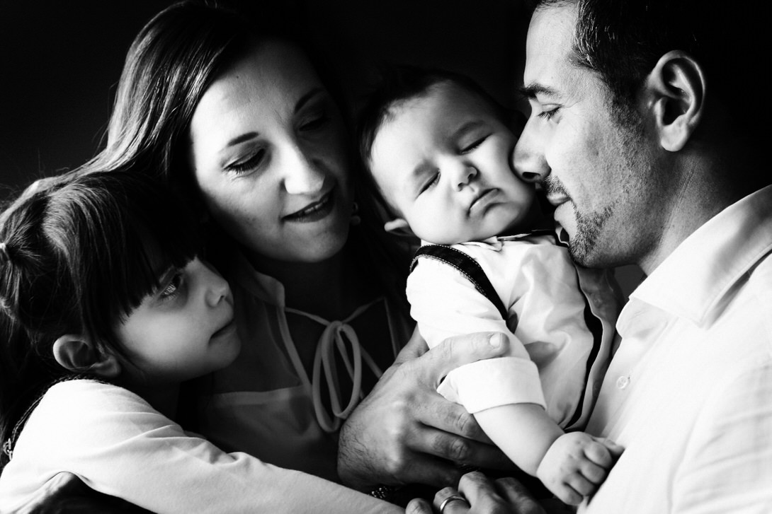 Famiglia felice in un ritratto in bianco e nero, con una madre che tiene un bambino addormentato e un padre che guarda affettuosamente la madre, mentre una bambina osserva con curiosità. L'immagine cattura un momento di tenerezza e connessione familiare.