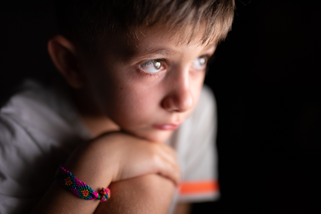 Ritratto di un bambino con sguardo pensieroso, appoggiato sul suo avambraccio, mentre indossa un braccialetto colorato. Lo sfondo scuro mette in risalto i suoi occhi chiari e l'espressione contemplativa.