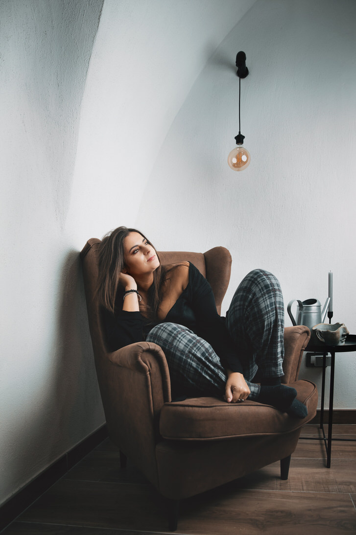 Donna seduta su una poltrona marrone, indossa un maglione nero e pantaloni a quadri, mentre guarda pensierosa verso l'alto. L'ambiente è luminoso, con pareti bianche e una lampada pendente sopra di lei. A fianco, un tavolino con accessori da tè e un vaso.