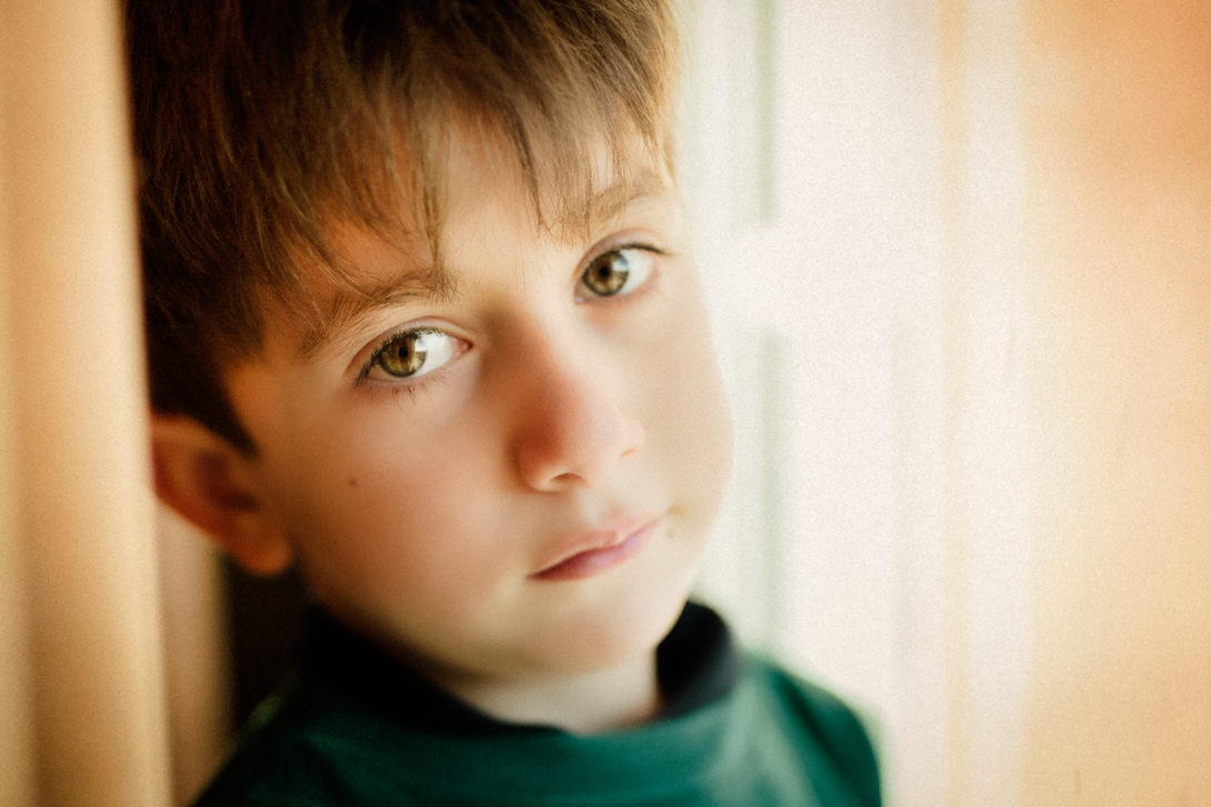Ritratto di un ragazzo con capelli castani e occhi chiari, in primo piano, mentre guarda verso l'obiettivo con un'espressione pensierosa. La luce soft si riflette sulla sua pelle, creando un'atmosfera calda e intima.