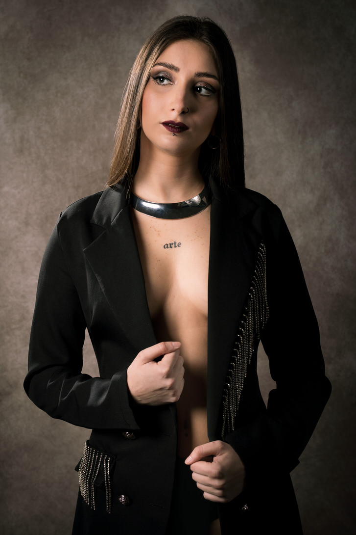 Modella in un abito elegante, con giacca nera e collana, posa con uno sguardo intenso su sfondo neutro. L'immagine mette in risalto la sua bellezza e sicurezza.