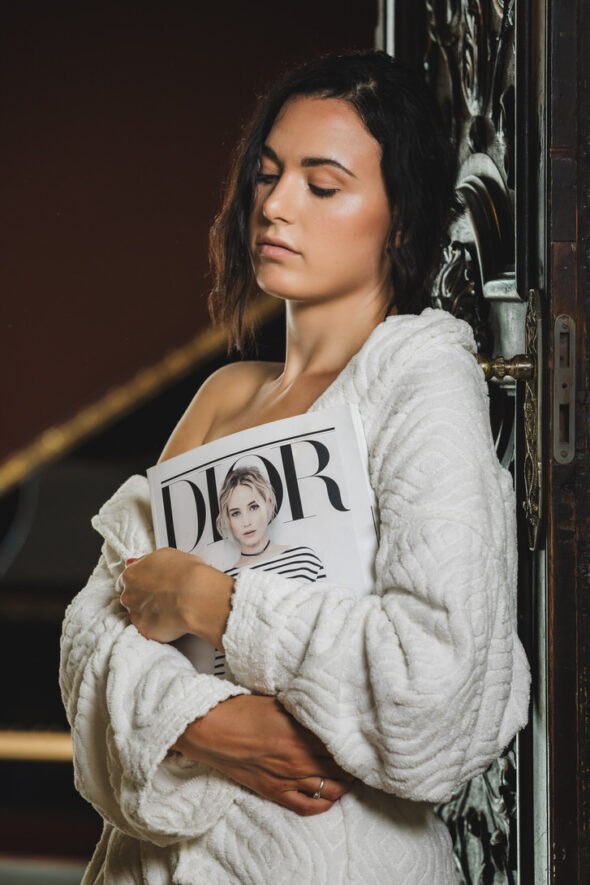 Modella in maglione bianco che abbraccia una rivista Dior, con espressione serena, in un ambiente elegante e raffinato.