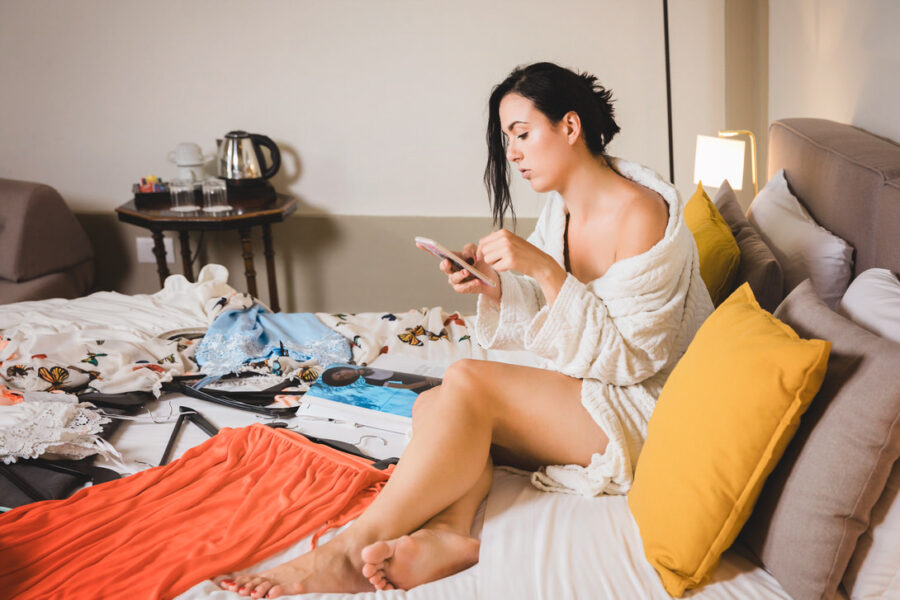 Donna seduta su un letto disordinato, indossa un accappatoio bianco e guarda il suo smartphone. Accanto a lei ci sono vestiti sparsi e accessori, creando un'atmosfera di relax e preparazione.