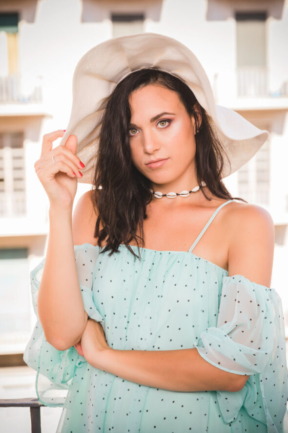 Modella con abito azzurro e cappello di paglia, posa in un ambiente luminoso con finestre sullo sfondo.