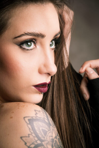 Ritratto di una giovane donna con occhi verdi e labbra scure, mentre tocca i suoi lunghi capelli castani. La pelle è decorata con un tatuaggio visibile sulla spalla. Lo sfondo è sfocato, evidenziando il suo sguardo intenso e il trucco elegante.