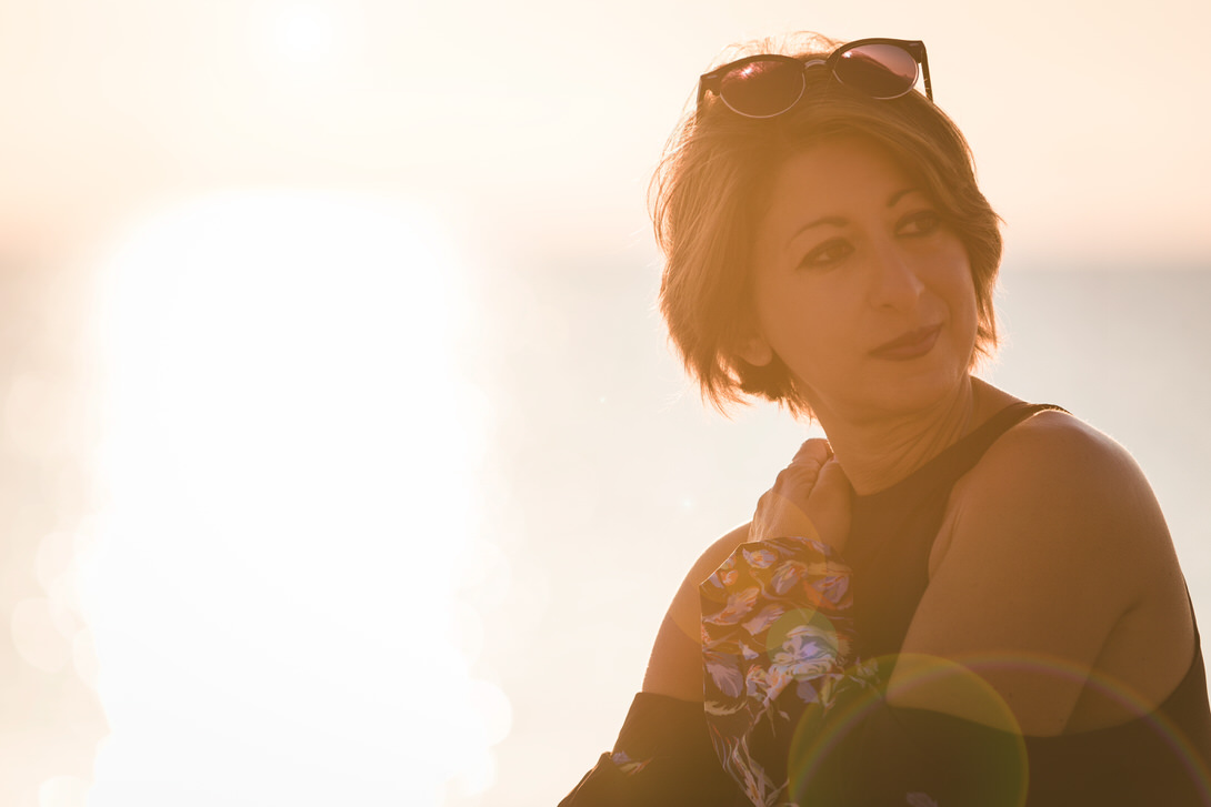 Donna con capelli corti e occhiali da sole, in posa contemplativa al tramonto, con uno sfondo luminoso che crea un'atmosfera serena.