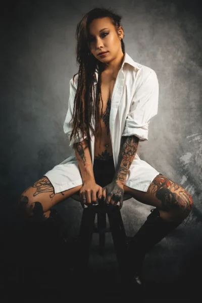 Modella con dreadlocks e numerosi tatuaggi, indossa una camicia bianca aperta e stivali neri, mentre posa seduta su uno sgabello con uno sfondo neutro. La foto evidenzia il suo stile audace e originale.