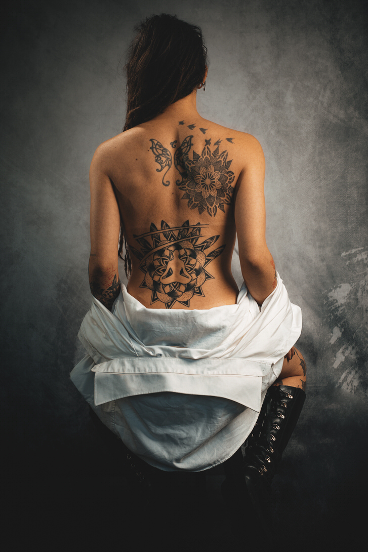 Modella con tatuaggi dettagliati sulla schiena posa con camicia oversize abbassata e stivali neri durante uno shooting fotografico artistico in studio