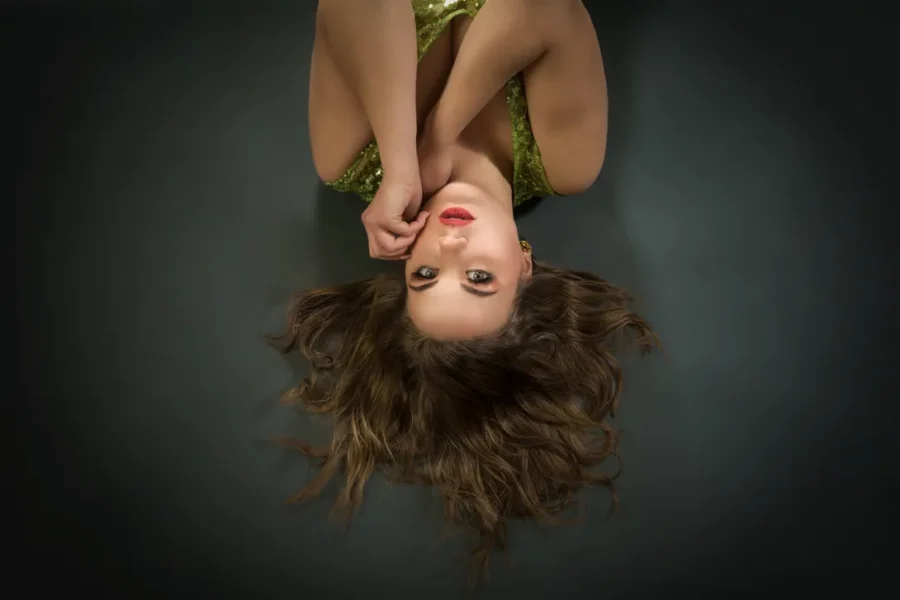 Ritratto di una giovane donna con capelli mossi, distesa su uno sfondo scuro. Indossa un abito verde brillante e posa con le mani sul viso, esprimendo un'aria di eleganza e mistero.