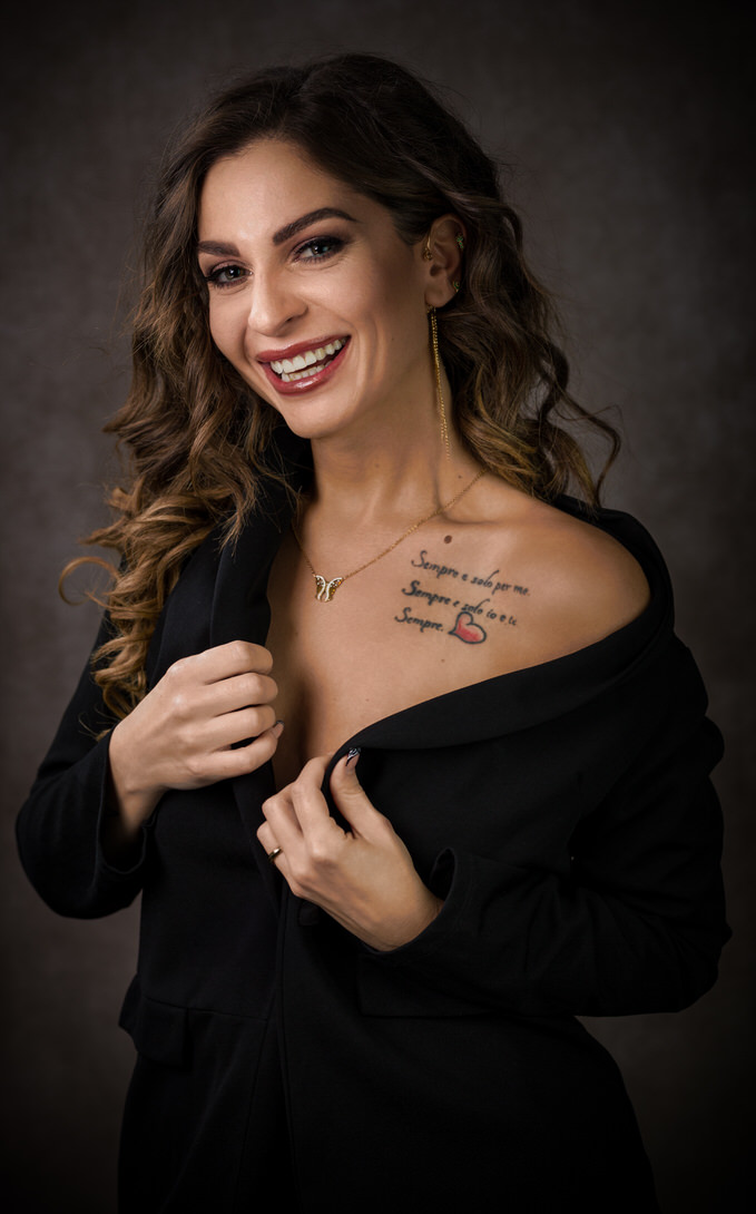 Ritratto di una giovane donna sorridente con capelli mossi, che indossa un abito nero. Mostra un tatuaggio sul petto con frasi significative e un cuore. La luce mette in risalto il suo sorriso e l'eleganza della sua posa.