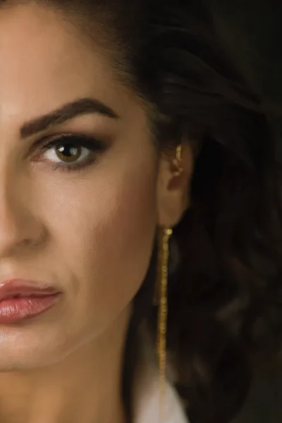 Primo piano di una donna con capelli ricci scuri, trucco elegante e orecchini dorati, che esprime un look raffinato e sofisticato.