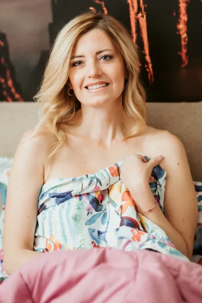 Donna sorridente con capelli biondi, avvolta in una coperta colorata, seduta su un letto. Lo sfondo presenta elementi artistici. L'immagine trasmette un'atmosfera di relax e comfort.