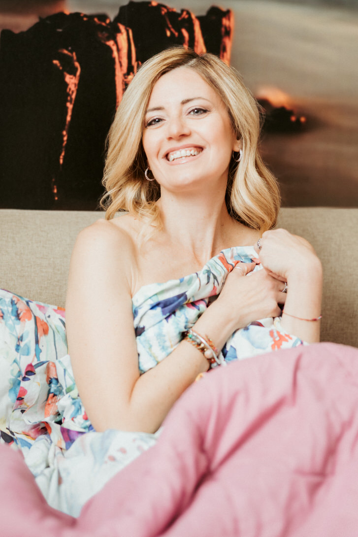 Donna sorridente avvolta in una coperta colorata, seduta su un divano. Lo sfondo mostra un paesaggio suggestivo. L'immagine trasmette un'atmosfera di comfort e serenità.