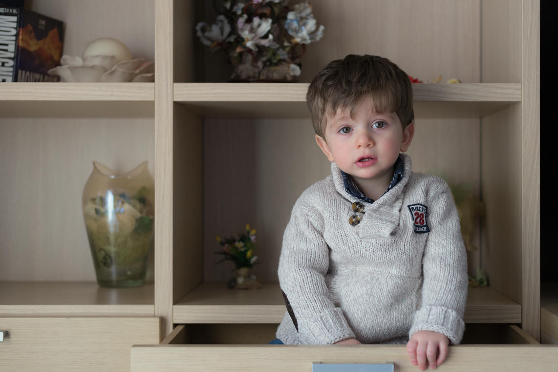 Bambino seduto in un cassetto di legno, indossa un maglione grigio chiaro e guarda verso la fotocamera. Sullo sfondo, una libreria con decorazioni e oggetti decorativi.