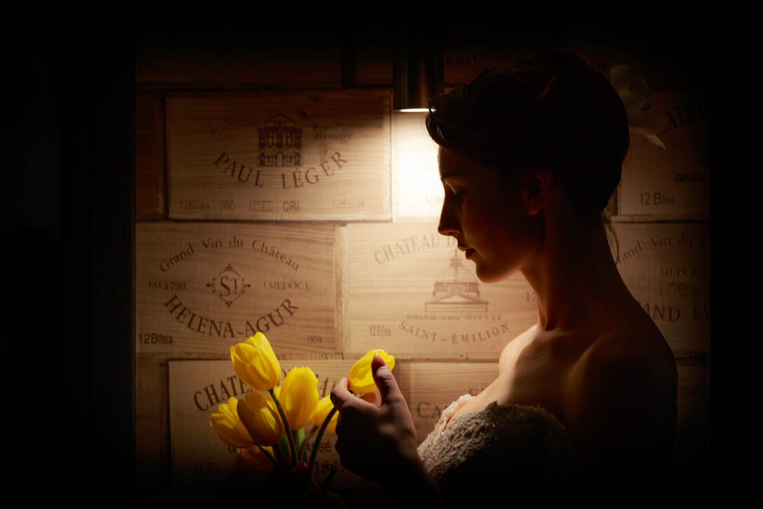 Workshop con Vittorio Lioce Ritratto di una donna con un bouquet di tulipani gialli, illuminata da una lampada, con sfondo di casse di legno di vino. La figura è in silhouette, evidenziando i dettagli del suo viso e dell'abito.