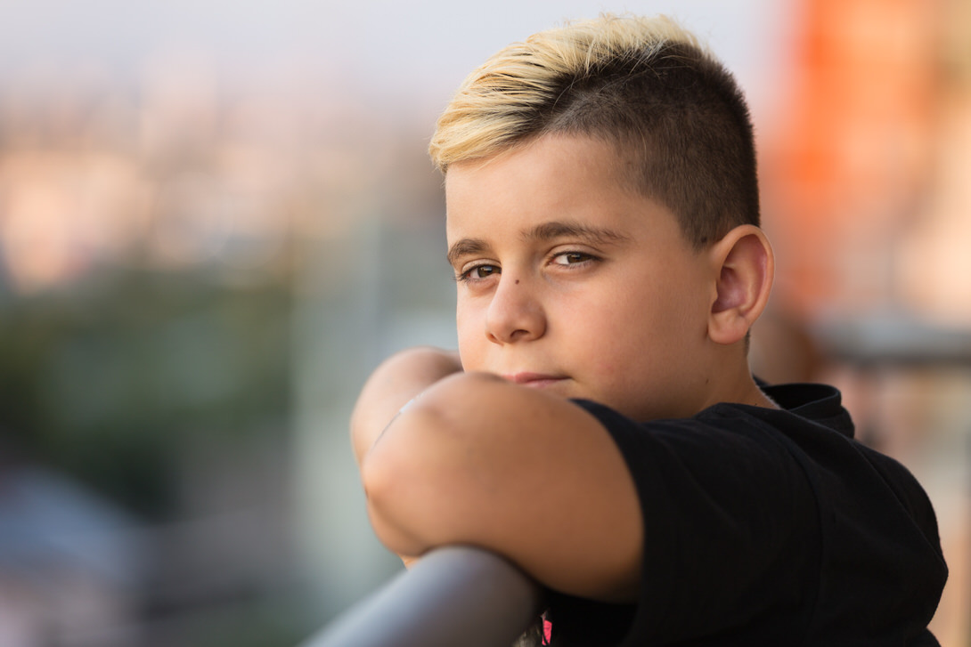 Ragazzo con capelli biondi e scuri che guarda in camera, appoggiato a un parapetto, con sfondo sfocato di una città.