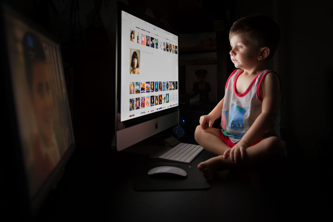 Bambino seduto davanti a un computer, concentrato su uno schermo luminoso che mostra una galleria di immagini. La scena è illuminata in modo soffuso, creando un'atmosfera tranquilla e coinvolgente.
