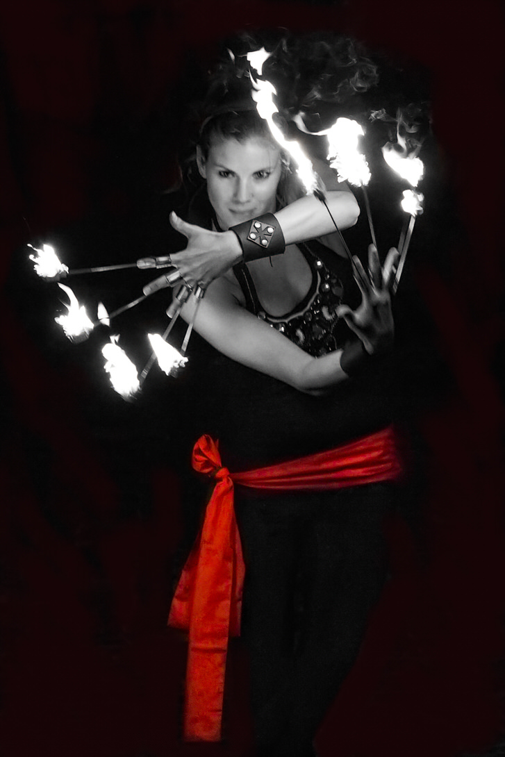 Lux Arcana Donna che esegue una performance di giocoleria con torce infuocate, indossando un top nero e un foulard rosso. Effetto artistico in bianco e nero con accenti di fuoco.