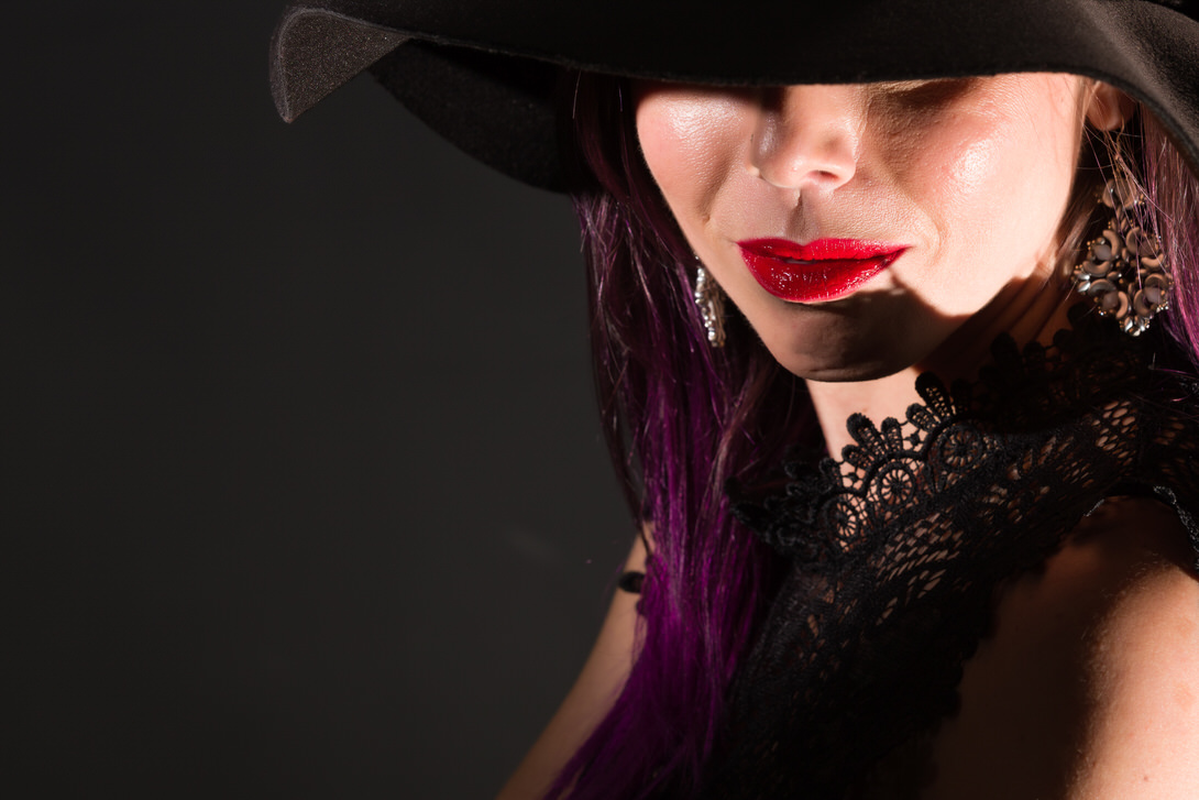 Ritratto di una donna con un cappello nero, labbra rosse e orecchini eleganti, in uno sfondo scuro che mette in risalto il suo trucco e i dettagli del suo abbigliamento.