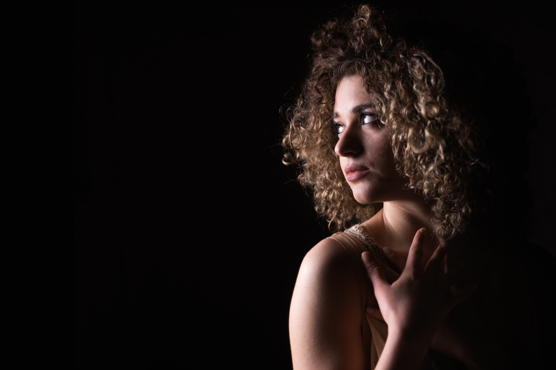 Ritratto di una giovane donna con capelli ricci, che guarda pensierosa verso sinistra, in un ambiente scuro, evidenziata da una luce soffusa che accentua i suoi lineamenti ed espressioni.