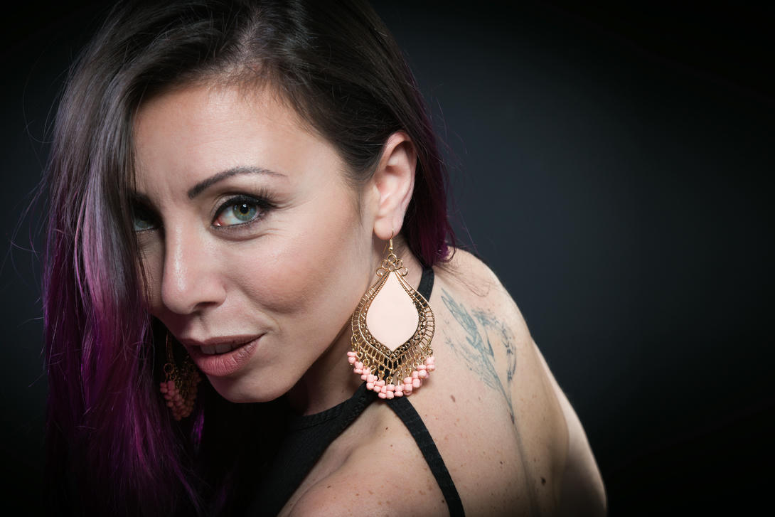 Antonella Rosati MUA Ritratto di una donna con orecchini pendenti eleganti e capelli neri con riflessi viola, che mostra un'espressione affascinante. Sullo sfondo scuro, si notano anche dei tatuaggi sulla spalla.