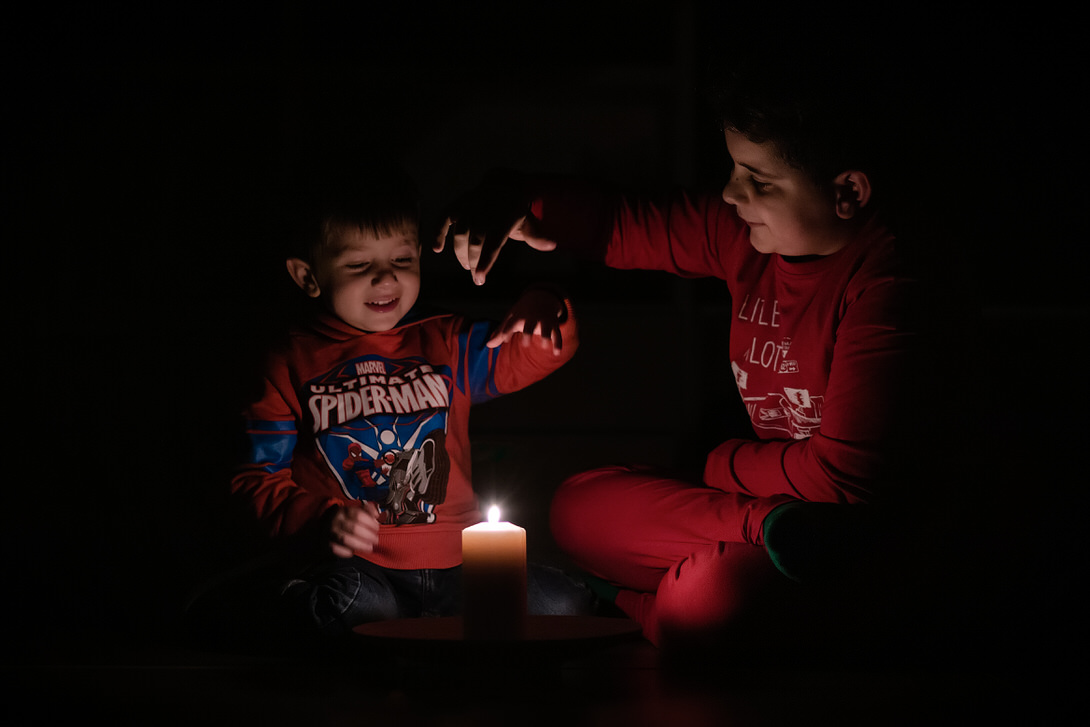 Due bambini sorridenti giocano con una candela accesa in un ambiente buio, creando un'atmosfera calda e accogliente. Il bambino a sinistra indossa una felpa di Spiderman, mentre il suo amico, a destra, è vestito di rosso.