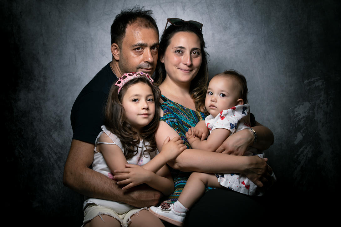 Ritratto di una famiglia composta da quattro membri, inclusi due bambini, abbracciati insieme in uno studio fotografico con sfondo grigio. La madre indossa una maglietta a fantasia colorata, mentre il padre è in camicia nera. Le bambine mostrano sorrisi estroversi, creando un'atmosfera di amore e unità familiare.