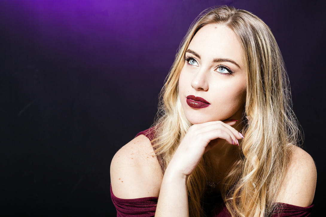 Ritratto di una giovane donna con capelli biondi lunghi e ondulati, che indossa un top scuro. Ha labbra rosse e guarda verso l'alto, pensierosa, con uno sfondo scuro e accenti viola.