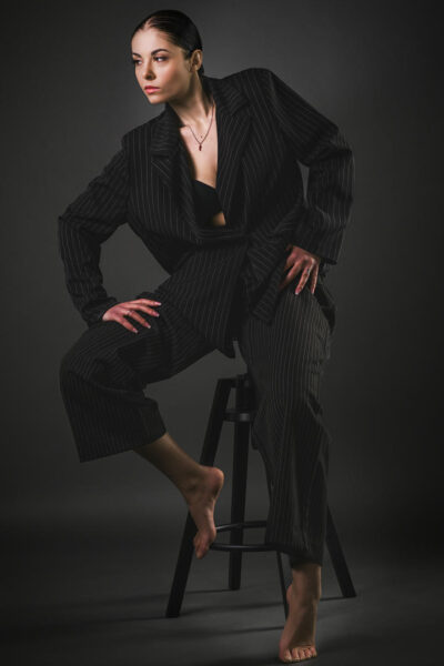 Modella in abbigliamento elegante a righe, posata su uno sgabello, con espressione seria e postura dinamica, su sfondo scuro.