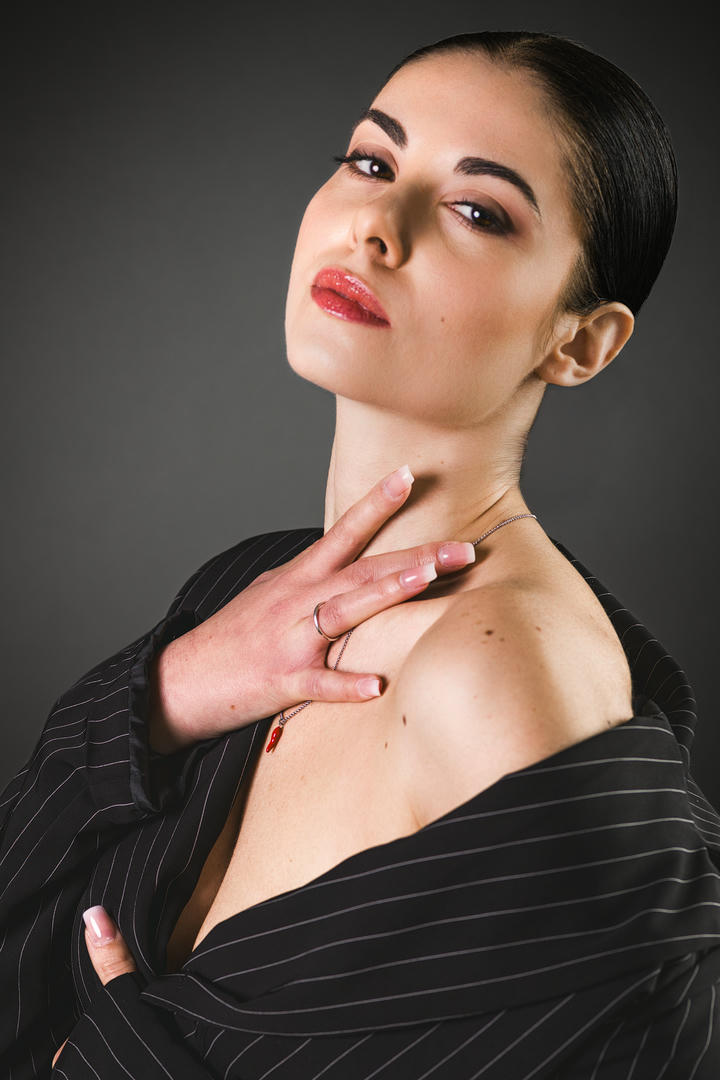 Donna elegante con trucco raffinato, indossa una camicia a righe e posa con grazia, evidenziando il collo e un gioiello delicato. Fondo scuro che mette in risalto il suo volto e la postura sofisticata.