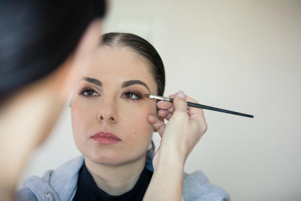 Trucco occhi professionale: una donna con la pelle chiara e capelli scuri riceve un'applicazione di ombretto da un truccatore, utilizzando un pennello.