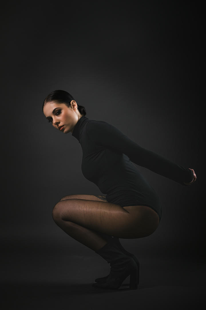 Modella in un abito nero attillato, acquattata su uno sfondo scuro, con calze e stivaletti neri. L'immagine enfatizza la sua postura e il trucco drammatico, creando un'atmosfera elegante e intensa.