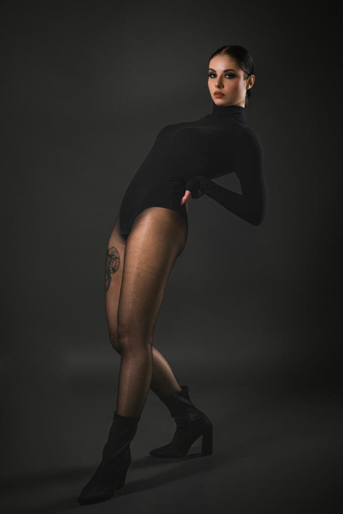 Modella in un bodysuit nero e calze trasparenti, posata in modo drammatico su sfondo scuro, evidenziando un tatuaggio sulla coscia e stivali alti.