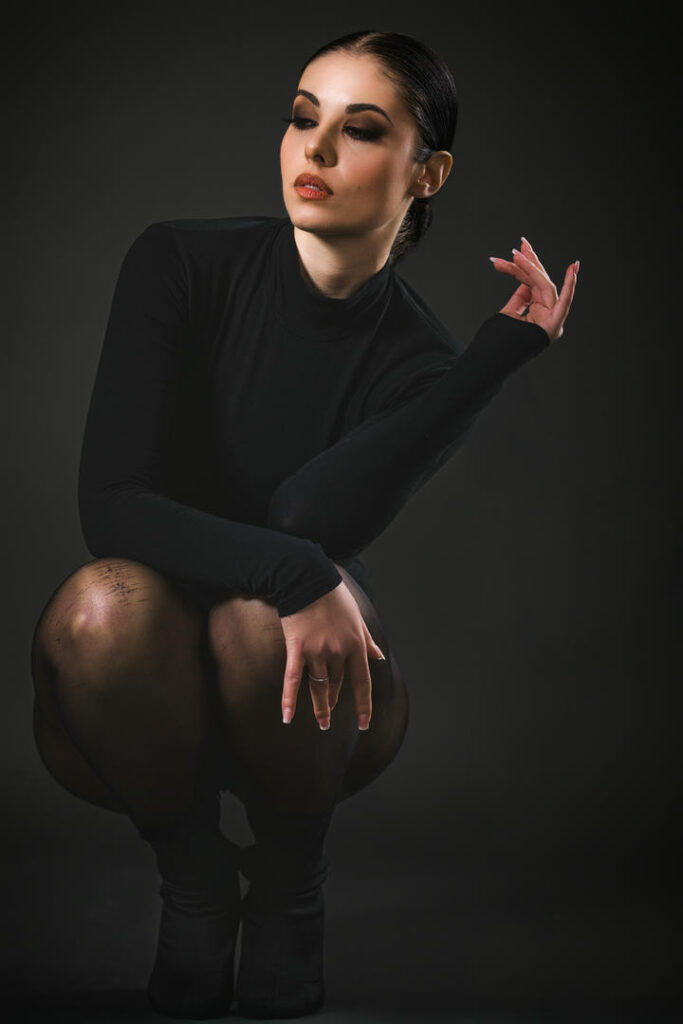 Modella professionale in posa elegante, vestita con un body nero e collant trasparenti, su sfondo scuro. La composizione evidenzia la sua espressione intensa e lo stile sofisticato.
