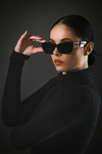 Modella con occhiali da sole neri in stile elegante, indossando un maglione a collo alto nero. Gli occhiali presentano dettagli dorati e sono posizionati con una mano in un pose sofisticata su sfondo scuro.