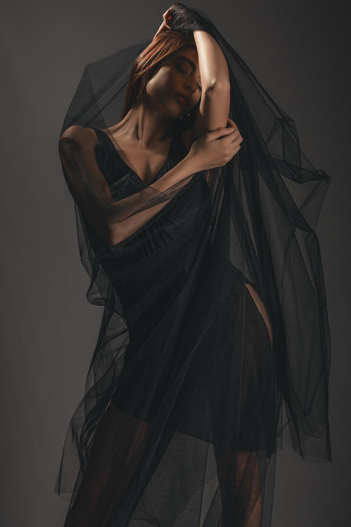 Ritratto in studio di Natalia Evangelista avvolta da un velo nero, illuminata lateralmente su fondale neutro, con una posa raccolta e uno sguardo abbassato che suggerisce una sensualità trattenuta e silenziosa.
