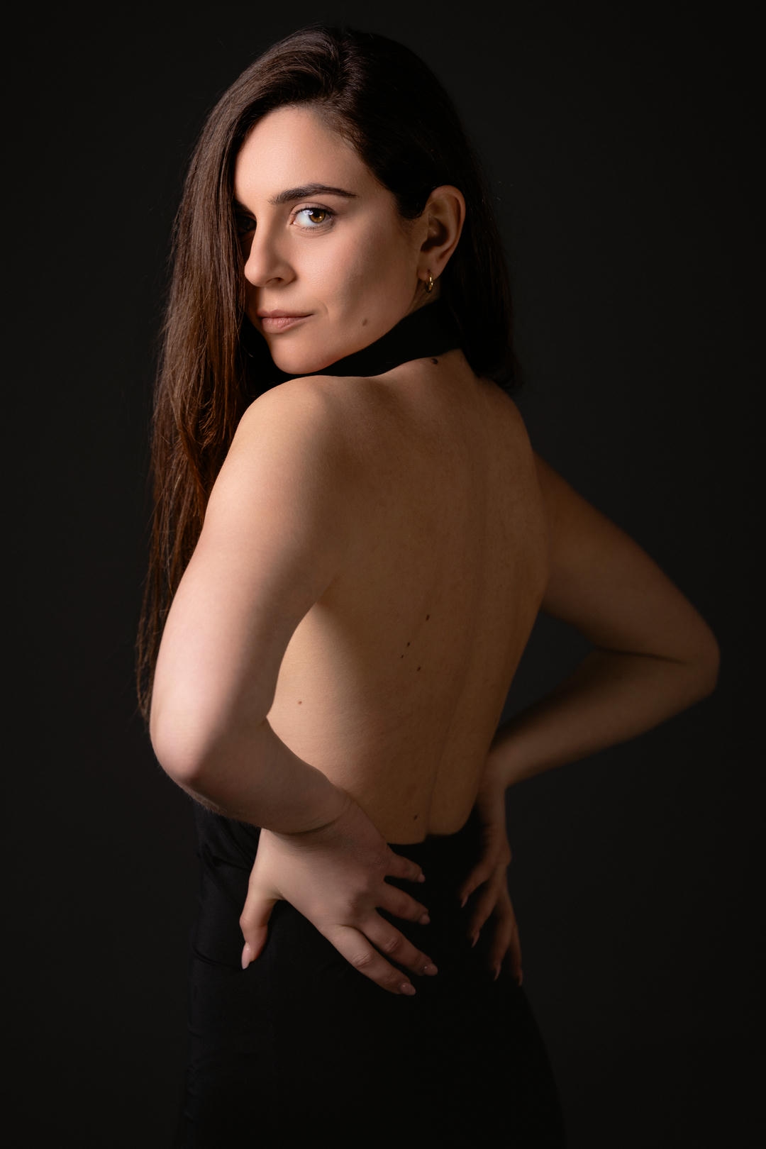 Ritratto femminile in studio con luce laterale su fondo scuro, figura in posa essenziale con schiena scoperta, fotografia minimalista centrata su presenza e controllo del corpo