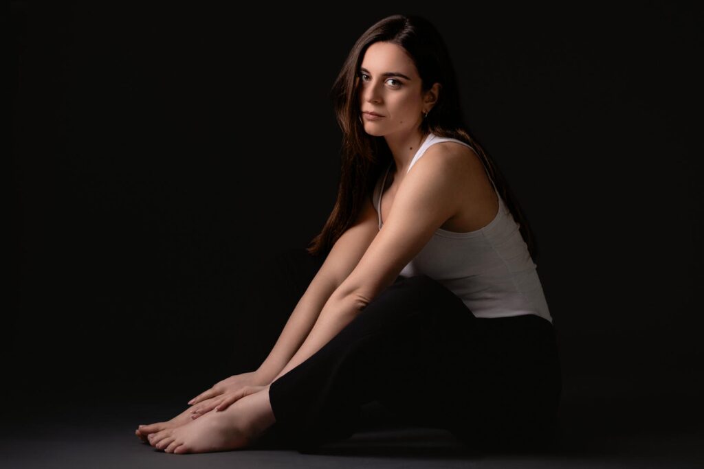 Ritratto fotografico in studio dal progetto “Senza far rumore”: figura femminile su fondo neutro scuro, luce laterale morbida che scolpisce volto e corpo, postura controllata e tensione trattenuta, espressione essenziale e non costruita, fotografia minimalista basata su presenza, sottrazione e controllo del gesto.